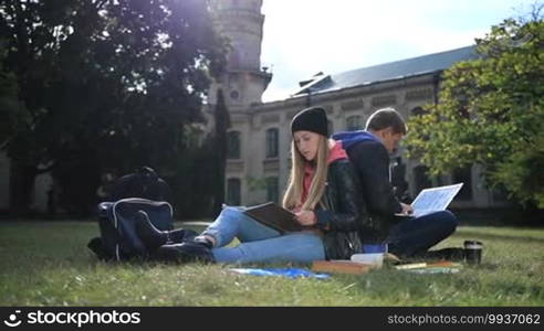 Young student couple sitting back to back on the lawn in the park outside the university campus and studying. The charming female student is reading a book, and her boyfriend is learning using a laptop.