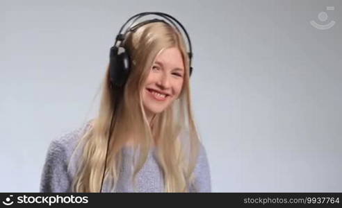 Young teenage woman listening to music intensely in her headphones over white background. Attractive girl hearing the music fading, touching the earcup and slowly taking headphones off, hanging them around her neck and smiling into the camera.