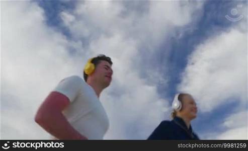 Young woman and man in headphones jogging against blue cloudy sky background. Everyday sports activity with music