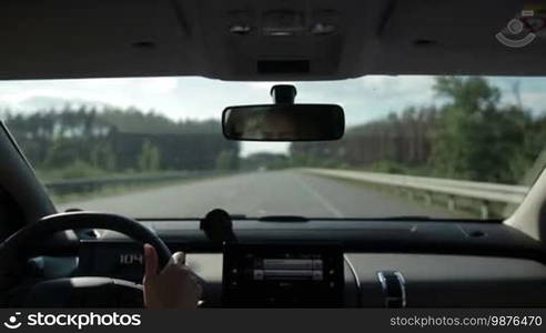 Young woman driving car on straight highway on sunny summer day. View from vehicle's interior. Female driver controlling auto on high speed on freeway.