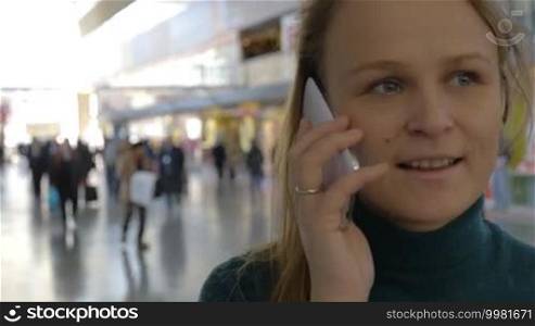Young woman having a phone talk in the station or airport hall. Defocused crowd of people in background