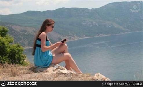 Young woman in a short blue dress reading the e-book by the sea