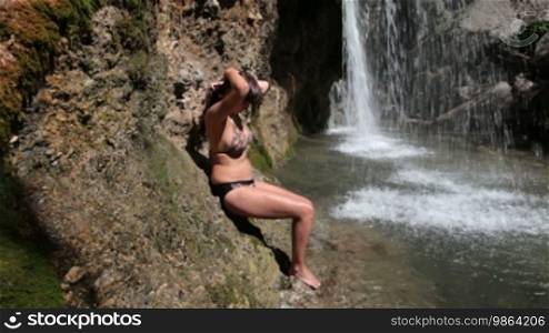 Young woman in bikini washes her face near the waterfall