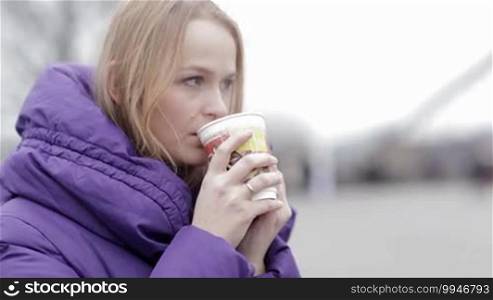 Young woman in purple jacket eating fast food and drinking hot tea outdoor in cold weather.