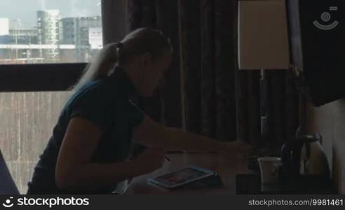 Young woman is working at table using tablet PC. She's turning the desk lamp on, answering the phone, and drinking beverage from the cup.