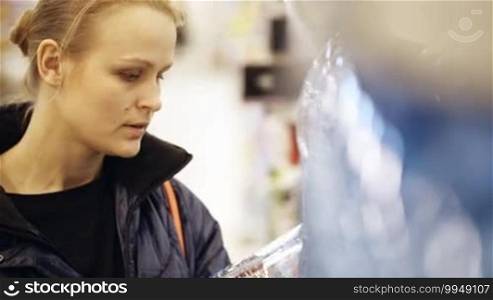 Young woman shopping in the supermarket. She is choosing a bottle of mineral water and putting it in her shopping basket