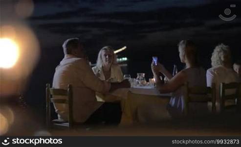 Young woman taking a picture of senior parents with a mobile phone during a late family dinner at the seaside. Defocused burning beach torch in the foreground