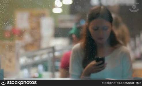 Young woman using cell phone to type text message or communicate in social media. She is sitting alone in the cafe in the evening, view through the wet glass