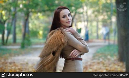 Young woman wearing fur vest, looking at camera, walking through the autumn park