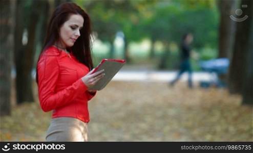 Young woman wearing red leather jacket with tablet computer in autumn park