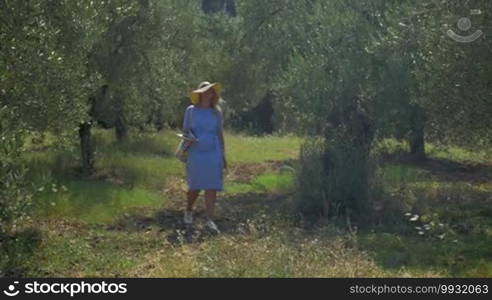 Young woman wearing summer hat wandering in olive orchard and taking photos of green nature with tablet computer