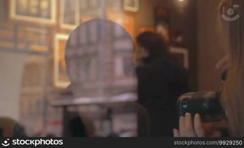 Young woman with digital camera taking shots of a boyfriend or friend in cafe, then they looking at photos on display. View through the window