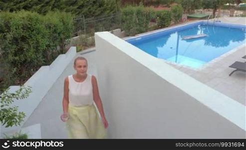 Young woman with glass of water walking from the yard with swimming pool to the house, she saying hello to man on the terrace and going indoor. Rent houses and villas for holidays