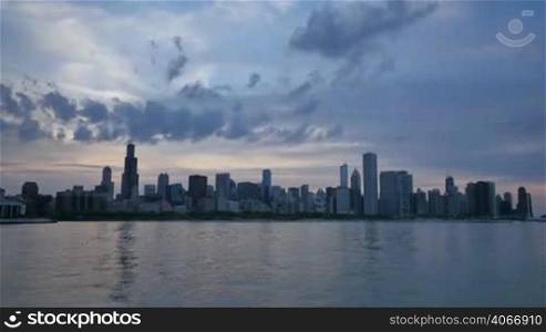 Video time lapse of Chicago downtown skyscrapers reflected on the Michigan lake from sunset clouds motion to night. Awesome Chicago city center skyline at night in the United States of America. Illuminated skyscrapers of Chicago in Spring twilight. Chicago sky in the evenig with the sun on the city sky.