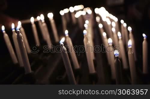 Viele Kerzen auf einem Kerzenstander brennen, auf der linken Seite nimmt eine Hand eine weitere Kerze und zundet sie an