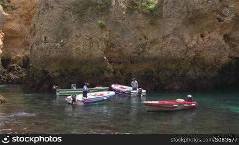 Vier Motorboote liegen vor einem gro?en sandsteinfarbenen Felsen im flachen turkisblauen Wasser, in zwei Booten stehen Manner. Kuste der Algarve, Portugal.