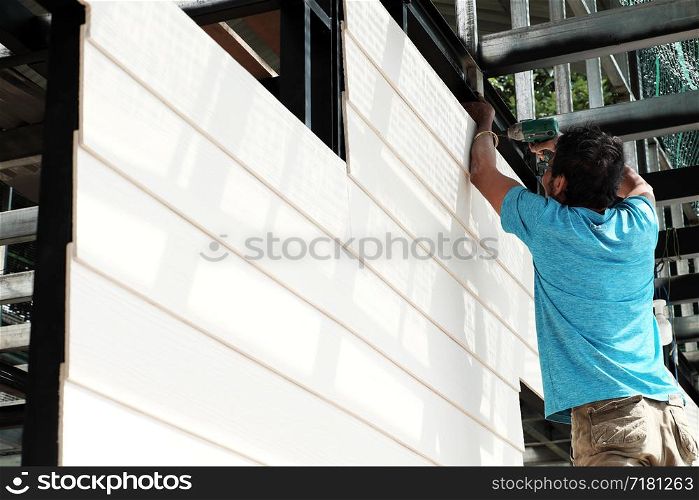 Vietnamese man stand on scaffold and shoot screw on smart wood panel to iron house frame, male do it yourself a house, Da Lat, Viet Nam