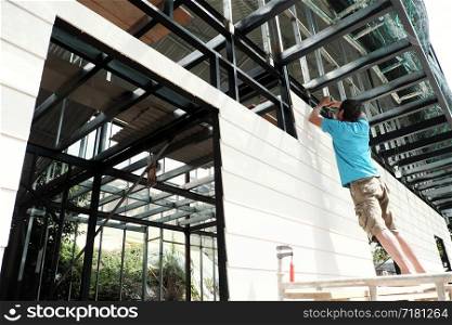 Vietnamese man stand on scaffold and shoot screw on smart wood panel to iron house frame, male do it yourself a house, Da Lat, Viet Nam