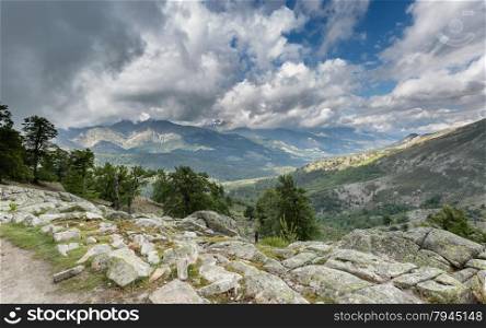 View across Corsican mountains from the GR20 track near to Lac de Nino with clouds and blue sky in the distance and trees and rocks in the foreground