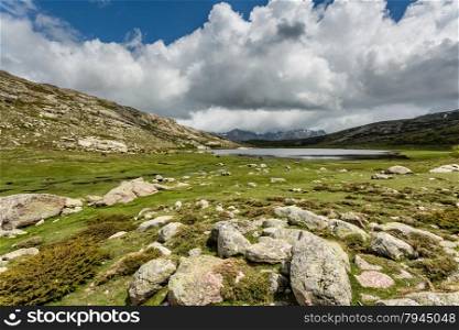 View across Lac De Nino in Corsica with a stream meandering across a green plain in foreground and dark clouds and snow capped mountains in the background