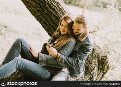 View at couple in love sitting on a tree in autumn park and using mobile phone