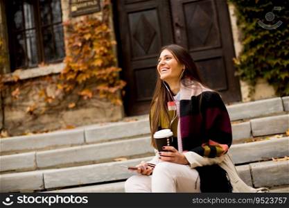 View at young woman sitting outside, drinking coffee to go and holding a mobile phone in hand