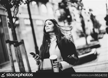 View at young woman standing outside, drinking coffee to go and holding a mobile phone in hand