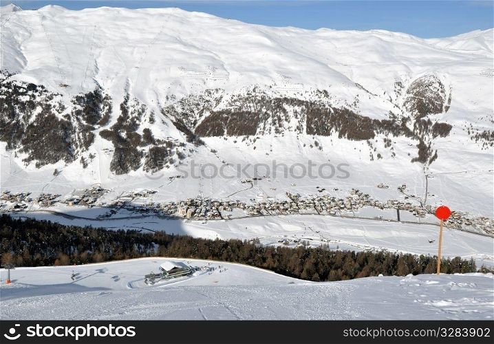 View down on lower station of modern chairlift with trees and a village down in the valley plus immense mountains in the background - shot in Livigno, Italian Alps