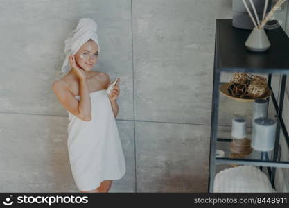 View from above of beautiful charming woman stands near grey wall in bathroom applies face lotion wrapped in white bath towel enjoys flawless of skin uses cosmetic product cares of complexion