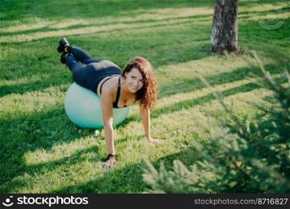 View from above of cheerful brunette woman makes pilates exercise with fitness ball, has sport training outdoor, poses on green grass in park, enjoys sunny day. Female performs yoga outside.