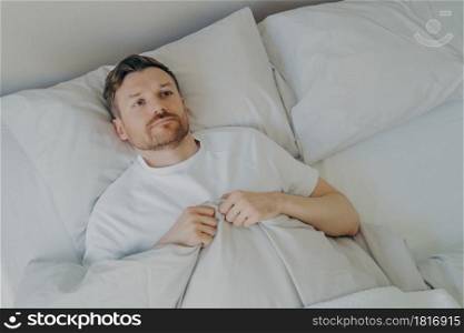 View from above of lonely upset young bearded man lying in bed with open eyes and can&rsquo;t sleep, feeling unhappy and tired, squeezes blanket with hands and being stressed of insomnia. Sleeping problems. Lonely upset young bearded man lying in bed with open eyes