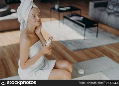 View from above of satisfied relaxed beautiful woman closes eyes and enjoys softness of skin, has beauty treatments after shower, holds bottle of lotion, poses at home, feels comfort and pleasure