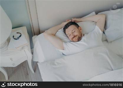 View from above of young happy relaxed caucasian man with attractive smile sleeping, lying in cozy bed with white beddings. Healthy care good night sleep and rest concept. Young happy relaxed caucasian man sleeping in bed