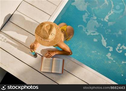 View from above on woman reading and drinking refreshing cocktail at swimming pool. Vacation and travel concept. View from above on woman reading and drinking at swimming pool