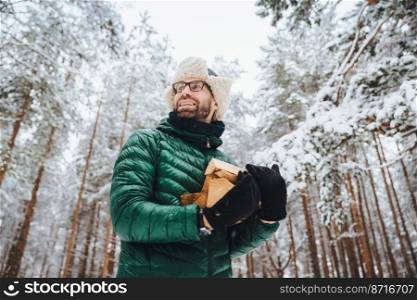 View from below holds firewood, looks happily aside, going to make fire, stands against beautiful white trees, admires majestic atmosphere in forest, spends weekends outdoors. People, winter concept