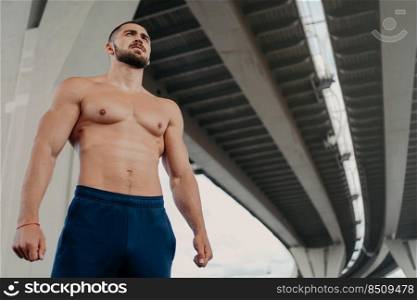View from bottom of self confident bearded handsome European man has strong perfect body looks into distance, leads active lifestyle, poses outside under bridge. Young shirtless bodybuilder.
