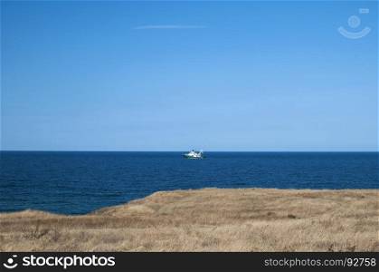 View from sea shore to calm blue sea with and fishing vessel on clear summer sky background