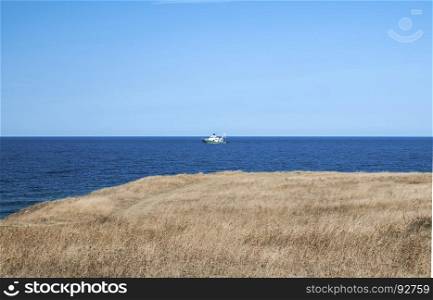 View from sea shore to calm blue sea with and fishing vessel on clear summer sky background