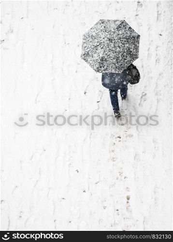 View from top at person walking on city streets during winter snowy weather. Person going through pavemet covered in white snow holding protective umbrella.. View from top at person with umbrella during winter