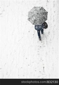 View from top at person walking on city streets during winter snowy weather. Person going through pavemet covered in white snow holding protective umbrella.. View from top at person with umbrella during winter