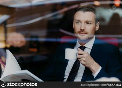 View from window of handsome successful young male boss, holds cup with hot aromatic beverage, looks through menu in luxury restaurant, dressed in formal expensive suit, thinks about something