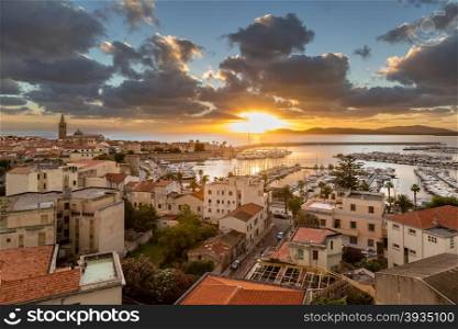 View looking over the city and harbour of Alghero on the west coast of Sardinia with the sun setting behind the lighthouse at Capo Caccia