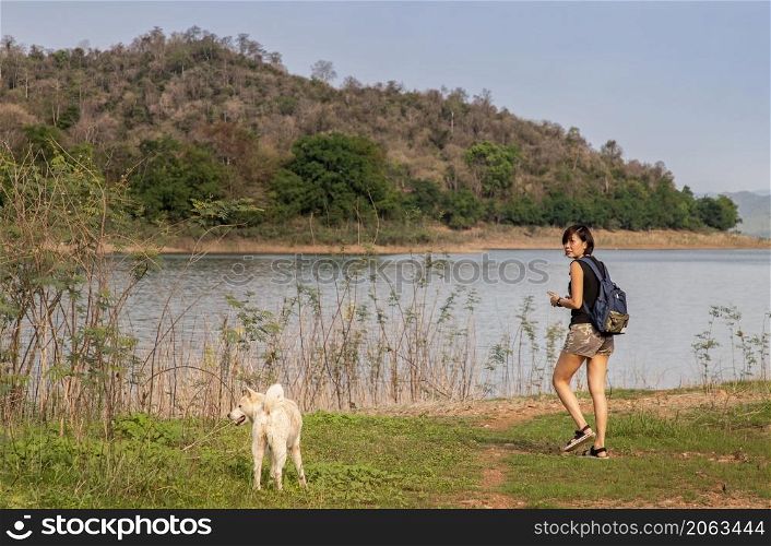 View of a young asia woman with white dog enjoying peaceful moment of beautiful view lake shore with mountains range in background. Pet and woman, Rest and enjoyment, lifestyle, Selective focus.