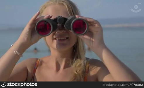 View of blond woman watching in binoculars with red glasses on landscape against blurred blue sea background