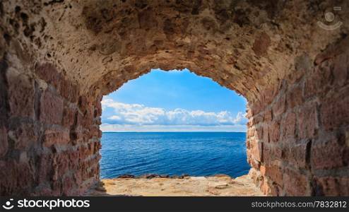 View of blue sea and sky from hole in old stonewall wall. Seascape in stone window casing frame. Travel tourism.