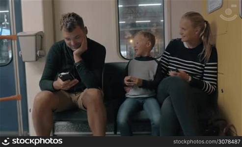 View of happy young family, mother, son and father, in the train on the stop station against window, sit on their seat places and watching smartphones and speaking. Amsterdam, Netherlands