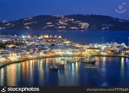 View of Mykonos Chora town Greek tourist holiday vacation destination with famous windmills, and port with boats and yachts illuminated in the evening blue hour . Mykonos, Cyclades islands, Greece. Mykonos island port with boats, Cyclades islands, Greece