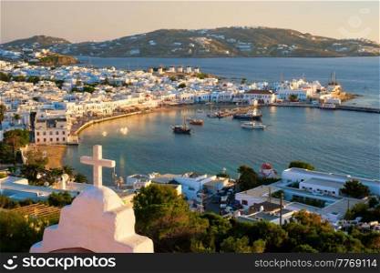View of Mykonos town Greek tourist holiday vacation destination with famous windmills, and port with boats and yachts on sunset over St Basil church cross. Mykonos, Cyclades islands, Greece.. Mykonos island port with boats, Cyclades islands, Greece