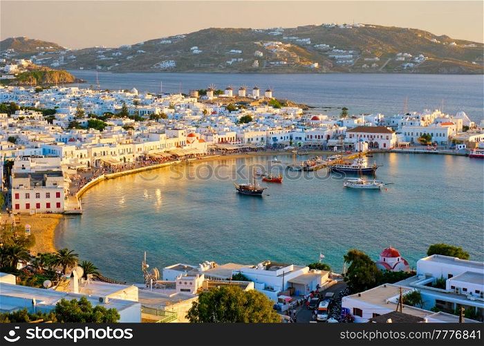 View of Mykonos town Greek tourist holiday vacation destination with famous windmills, and port with boats and yachtson sunset . Mykonos, Cyclades islands, Greece. Mykonos island port with boats, Cyclades islands, Greece