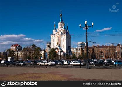 View of one of the streets of Khabarovsk in the daytime against a blue bright sky and white clouds. The combination of ancient architecture with modern buildings.. Khabarovsk, Russia - Apr, 27, 2019: Grado-Khabarovsk Cathedral of the Assumption of the Mother of God. Komsomolskaya square in the daytime with a bright blue sky with white clouds.